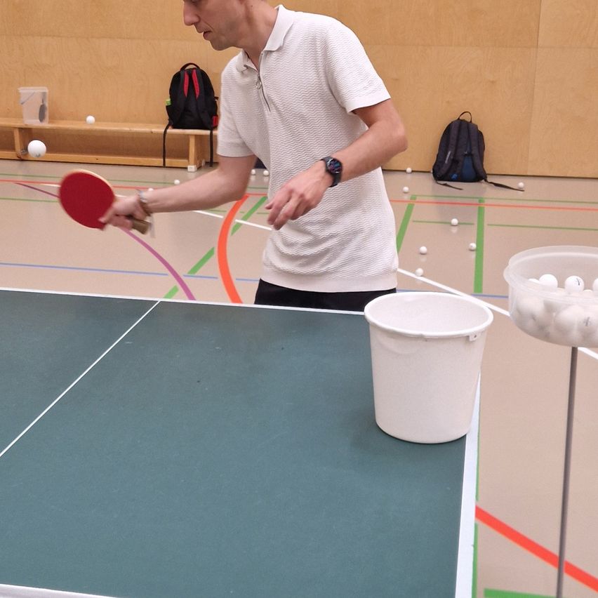 Ein Mann in einem weißen Hemd spielt Tischtennis mit einem roten Schläger in einem Fitnessstudio. Der Tischtennistisch ist blau und weiß. Ein Eimer mit Tischtennisbällen steht auf der rechten Seite.