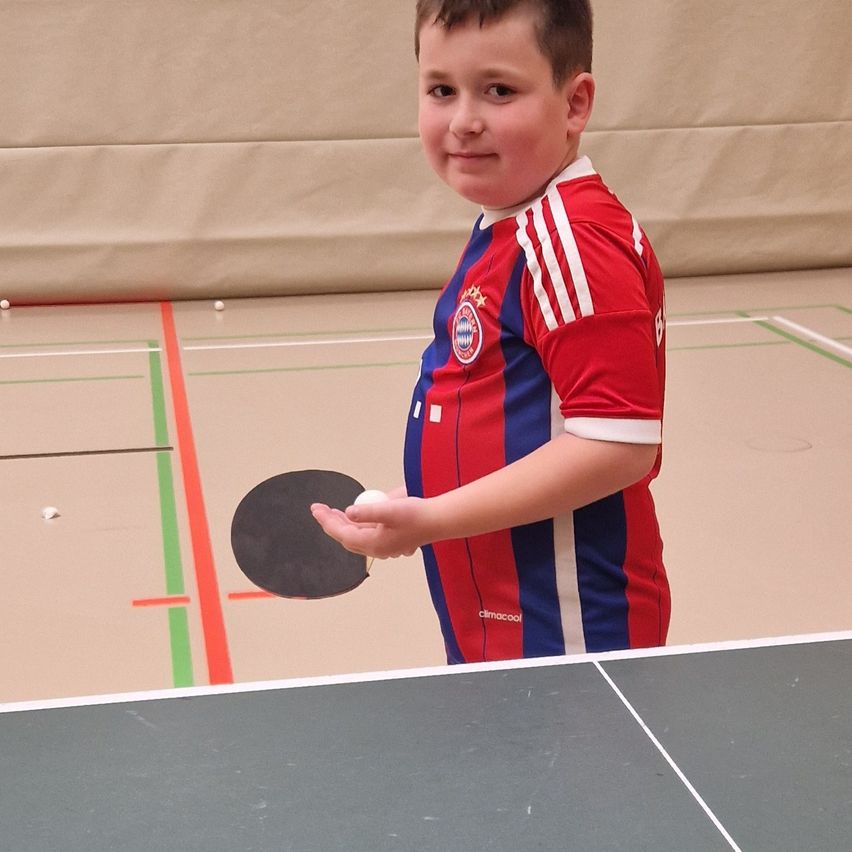 Ein Junge in einem rot-blau-weiß gestreiften Shirt steht in einer Tischtennishalle, hält eine Schläger und einen Ball.