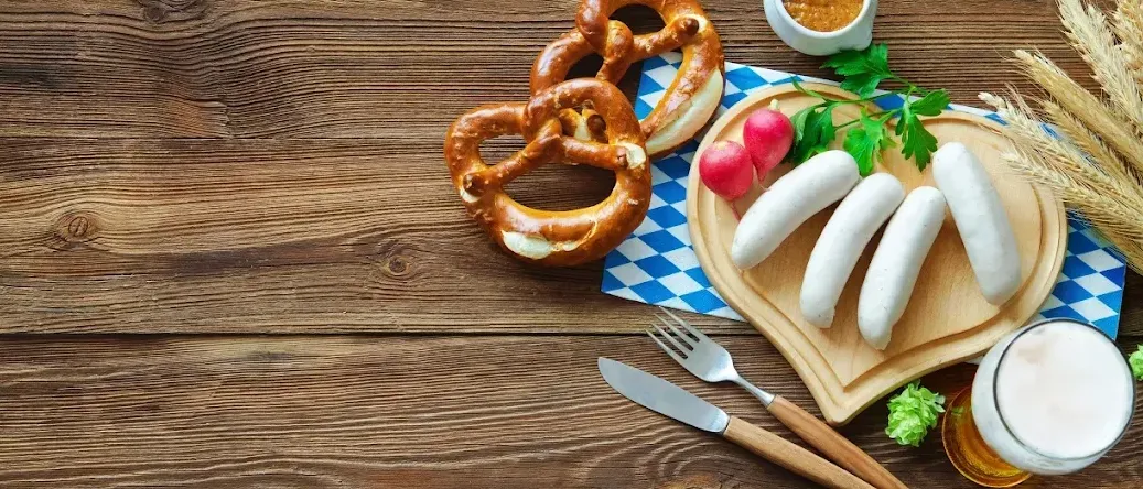 Ein hölzerner Tisch zeigt einen herzförmigen Teller mit Würsten, Radieschen und Petersilie. Zwei Brezeln liegen auf einem blau karierten Tuch. Ein Schüssel mit Senf und ein Löffel sind in der Nähe. Ein Messer und eine Gabel liegen auf dem Tisch.