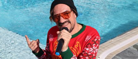 Ein Mann in einem roten Weihnachtspullover und einer schwarzen Mütze steht am Pool, hält ein Mikrofon in der Hand und lächelt.