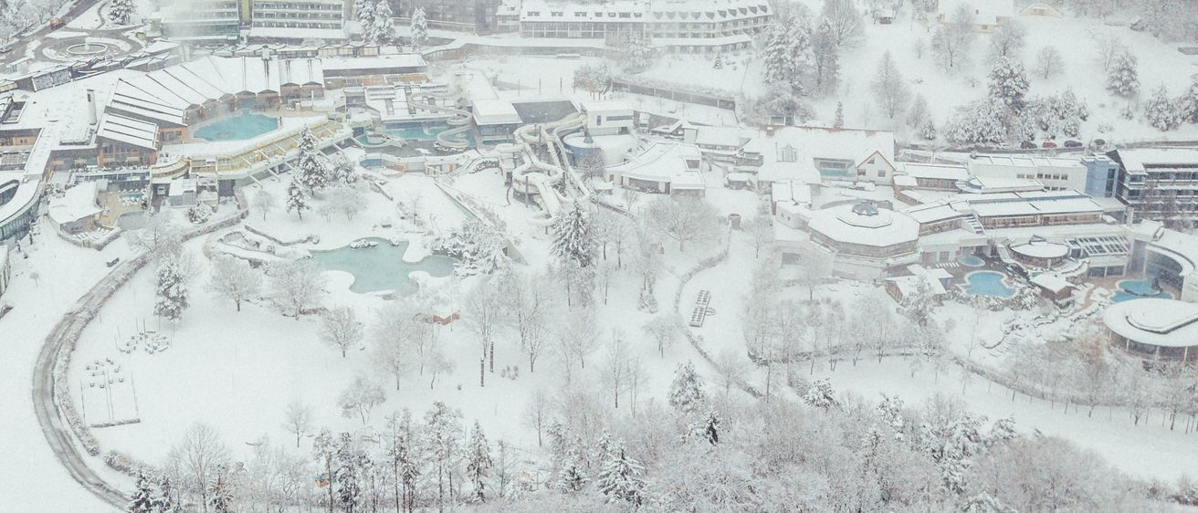 Luftaufnahme einer verschneiten Landschaft mit einem großen Resort, Schwimmbädern und Gebäuden, umgeben von schneebedeckten Bäumen und Hügeln.