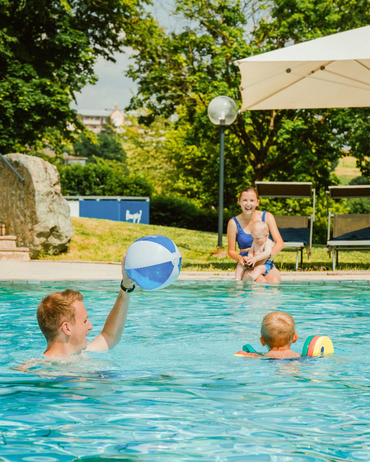 Eine Familie genießt einen Tag am Pool. Ein Mann hält einen Ball, eine Frau hält ein Baby und ein Kind schwimmt mit einem Schwimmhilfe.