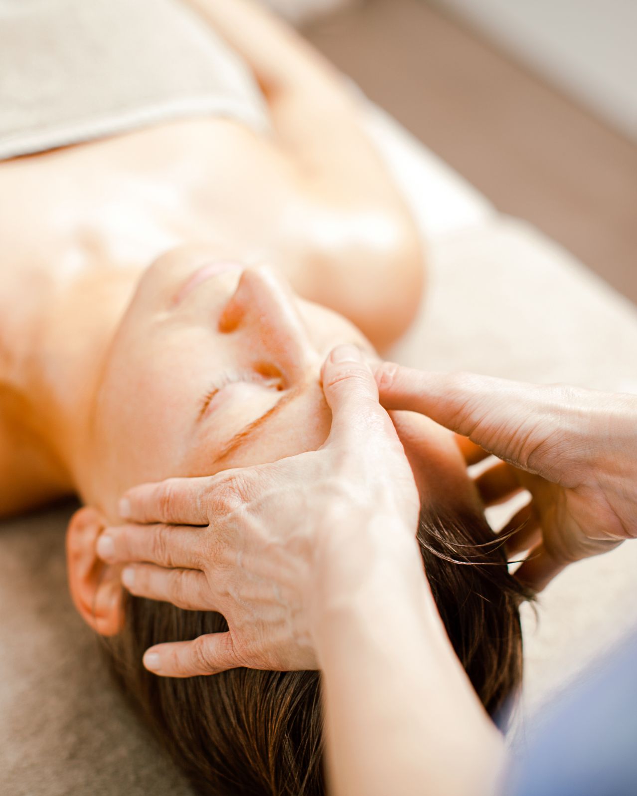 Eine Frau liegt auf einem Tisch und erhält eine Gesichtsmassage mit geschlossenen Augen, ihr Gesicht wird sanft von den Händen einer anderen Person gehalten.