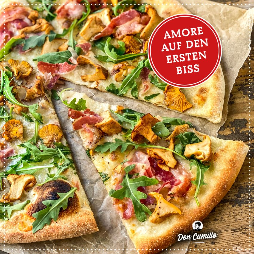 Zwei Pizzascheiben mit Pilzen, Speck und Rucola liegen auf Backpapier. Ein roter Kreis mit dem Text 'AMORE AUF DEN ERSTEN BISS' ist obenauf. Die Marke 'Don Camillo' ist unten sichtbar.