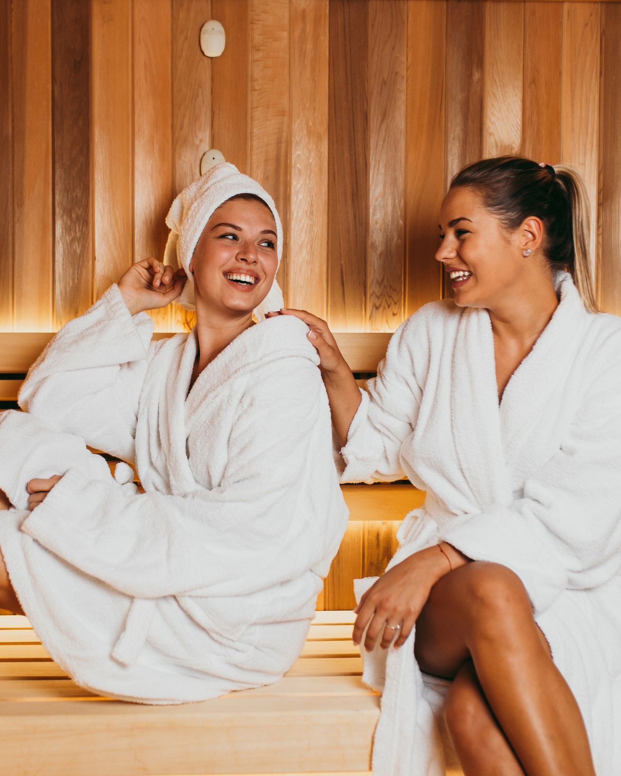 Zwei Frauen lächeln und sitzen auf einer Holzbank in einer Sauna. Sie tragen beide weiße Bademäntel.