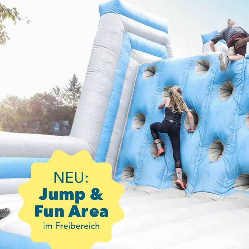 Eine aufblasbare blaue Struktur mit Löchern und der Aufschrift 'NEU: Jump & Fun Area im Freibereich'. Zwei Kinder klettern darauf.