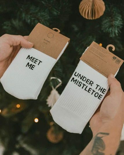 Two hands hold up two socks in front of a Christmas tree. The socks have printed text: one says 'Meet Me' and the other says 'Under the Mistletoe'.