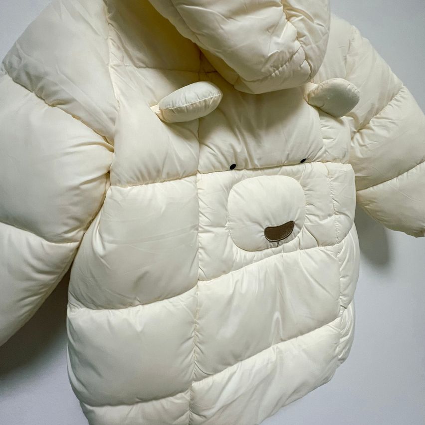 A white puffy jacket with a bear face on the front and hood, hanging on a wall.