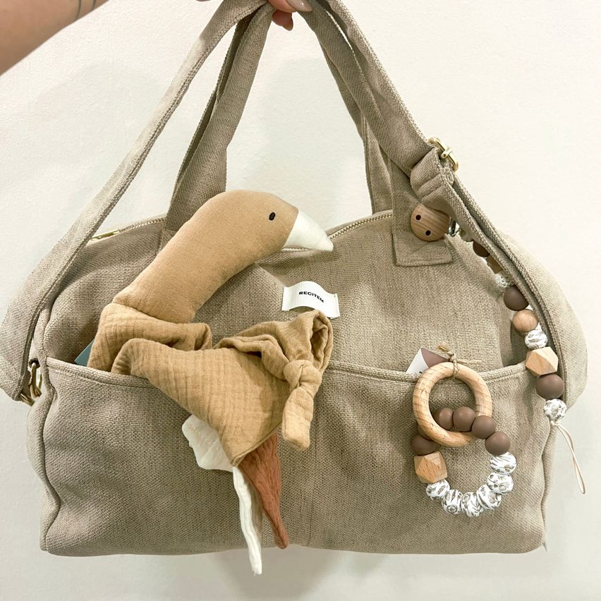A beige canvas tote bag with a duck toy and wooden beads inside. The bag has a handle and a zipper. The duck toy is folded in the bag's pocket.