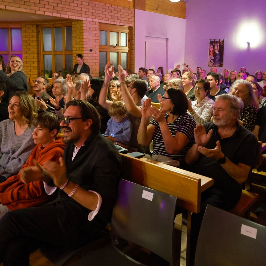 Eine große Gruppe von Menschen sitzt in einem Auditorium, klatscht und lächelt, wobei einige ihre Hände heben. Der Raum hat Ziegelwände und ein lila Licht.