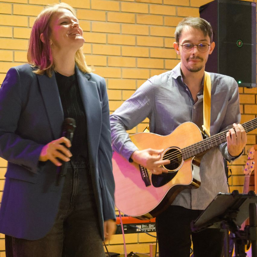 Eine Frau mit rosa Haaren steht und singt, während ein Mann eine Gitarre in einem Raum mit Ziegelwänden spielt.