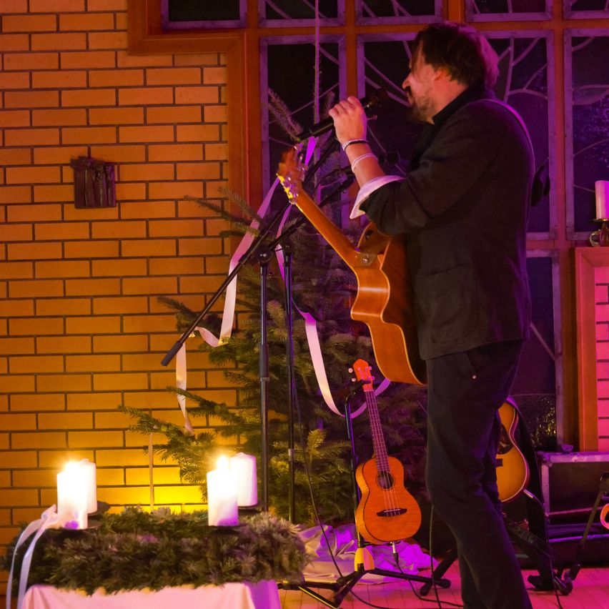 Ein Mann singt in ein Mikrofon, während er eine Gitarre auf einer Bühne spielt, mit einem Weihnachtsbaum, Kerzen und einer Ziegelwand im Hintergrund.