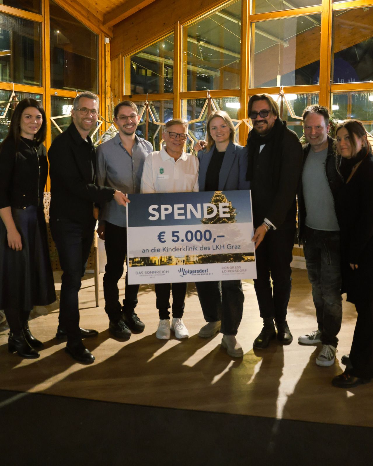 Eine Gruppe von Menschen steht zusammen und hält ein Schild. Es steht 'Spende' und '5.000 EUR'. Sie lächeln und scheinen für ein Foto zu posieren.