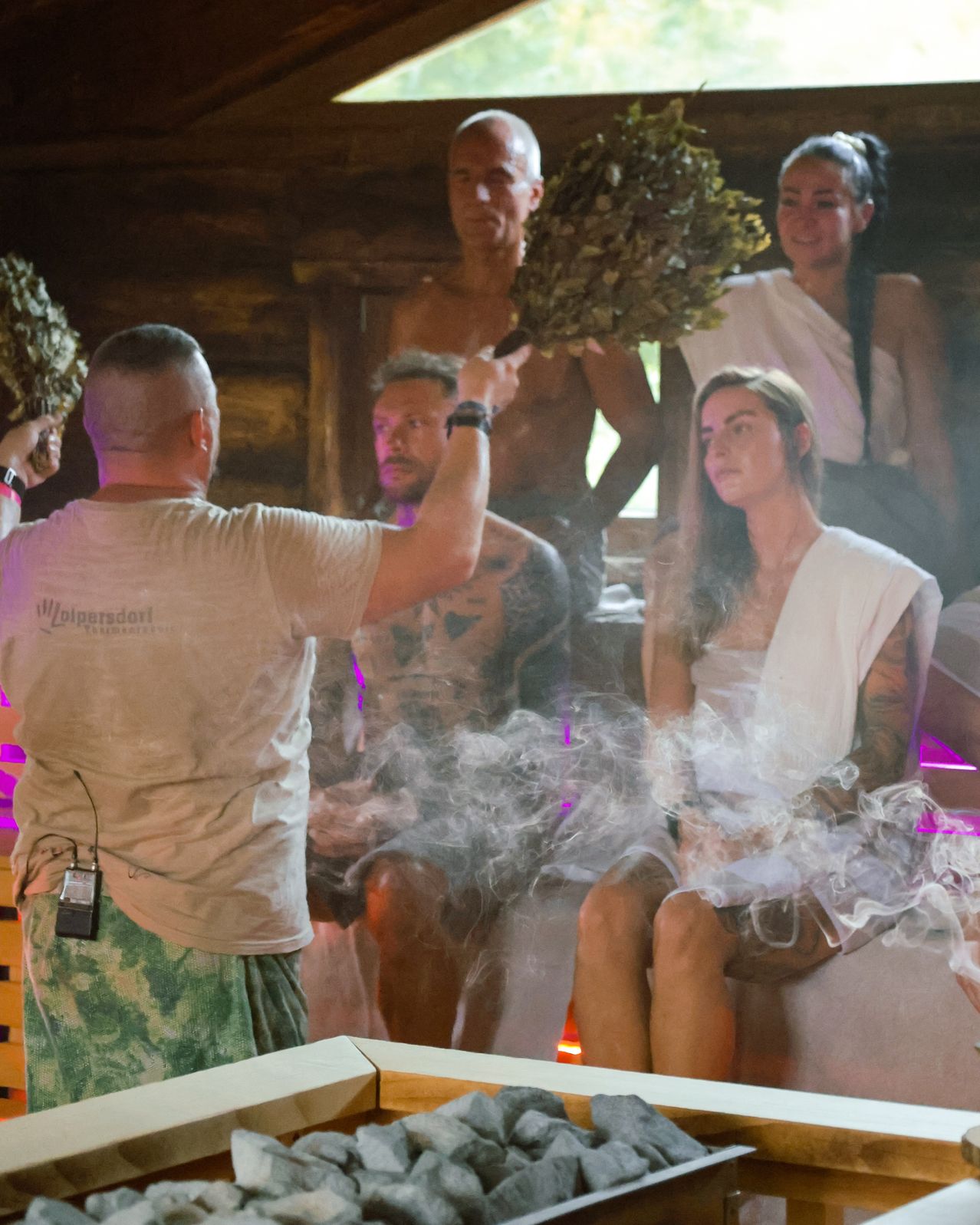 Eine Gruppe von Menschen sitzt in einer Sauna. Ein Mann steht vor ihnen, hält eine Pflanze und ein Mikrofon. Die Menschen tragen weiße Tücher. Der Mann trägt ein weißes Hemd und grüne Shorts. Das Licht reflektiert auf den Menschen.