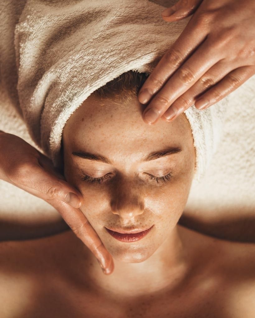 Eine Frau mit einem Handtuch auf dem Kopf erhält eine Gesichtsmassage, die Augen geschlossen und den Kopf leicht nach hinten geneigt.