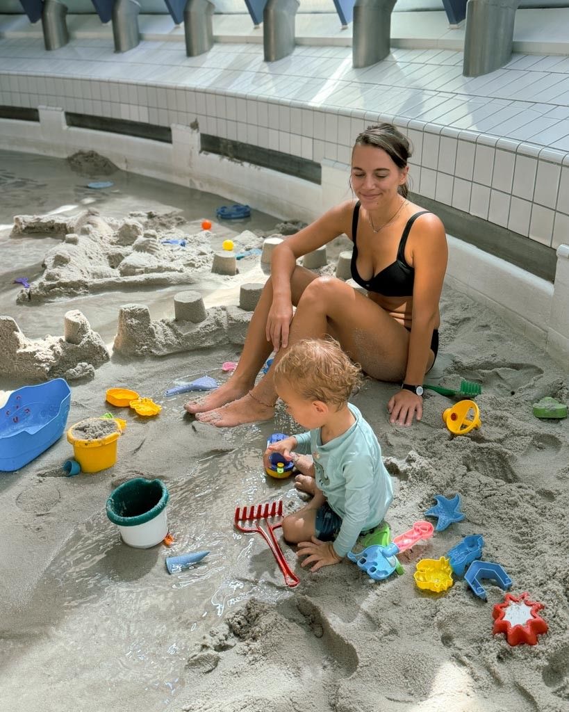 A woman and a young boy play in a sandpit with various toys. The woman is sitting on the sand while the boy is kneeling and holding a toy.