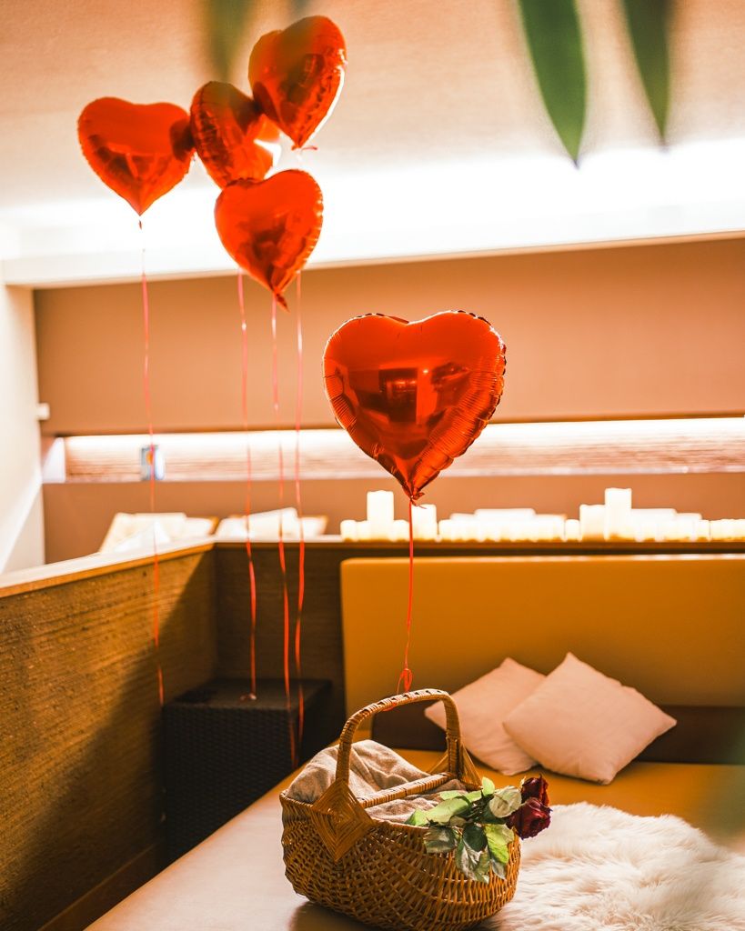 A cozy room with a beige sofa adorned with heart-shaped balloons and a wicker basket containing a bouquet of roses. Two white pillows are placed on the sofa, and a candle-lit area is in the background.