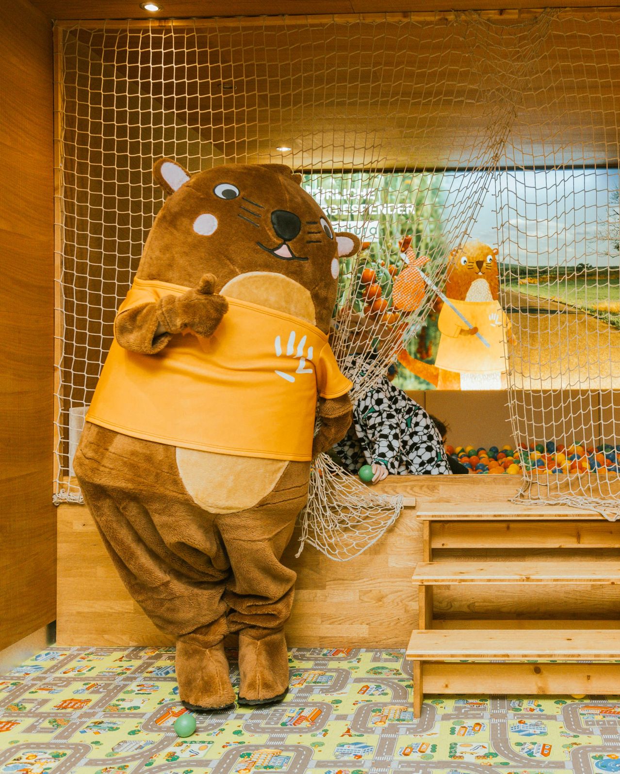 Ein Maskottchen, das als Biber verkleidet ist und ein gelbes Shirt trägt, steht in einem Spielbereich mit einem Netz und einem großen Bildschirm im Hintergrund. Ein Kind sitzt auf dem Boden und spielt mit Bällen.