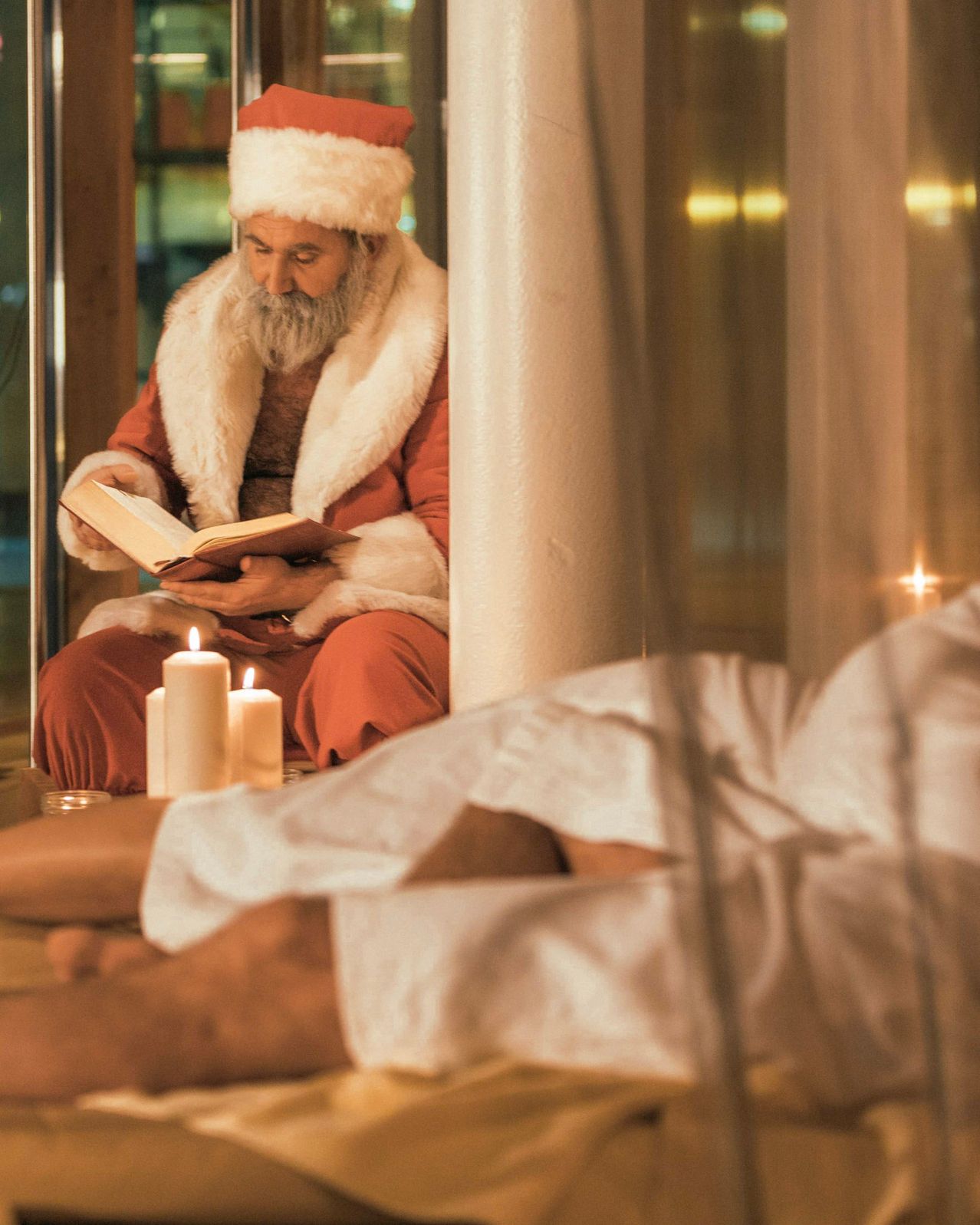 Der Weihnachtsmann in seinem traditionellen roten Kostüm sitzt neben einer Kerze und liest ein Buch. Ein Mensch liegt neben ihm in einem Bademantel.