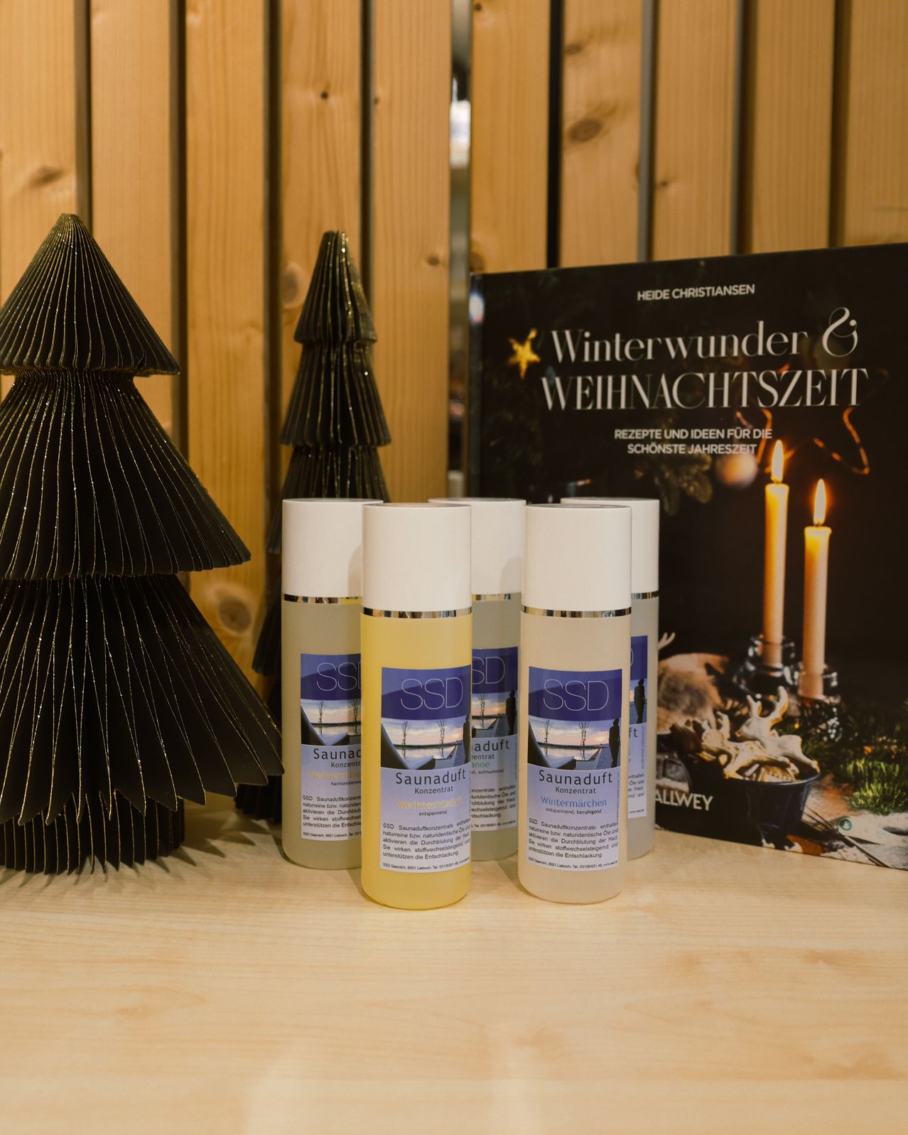 Auf einem Holztisch steht ein Buch mit dem Titel 'Winterwunder & Weihnachtszeit', Kerzen, ein schwarzer Weihnachtsbaum und vier Flaschen Saunaöl mit der Aufschrift 'SSD'.