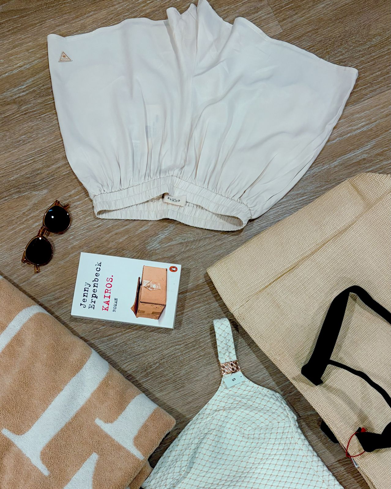 Ein weißes Kleidungsstück, eine Sonnenbrille, ein Buch mit dem Titel Kairos, ein Handtuch mit einem J-Monogramm, ein Netz-BH und eine beige Tasche liegen auf einer Holzoberfläche.