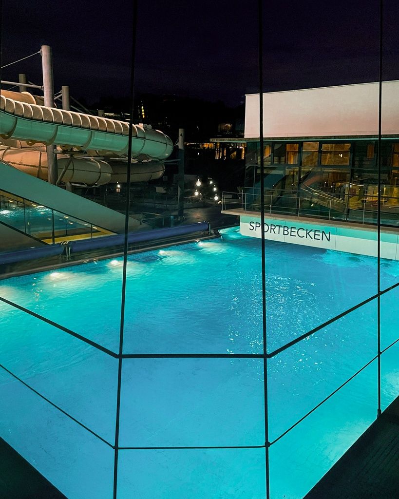 An illuminated swimming pool at night with a slide and a building labeled Sportbecken in the background.
