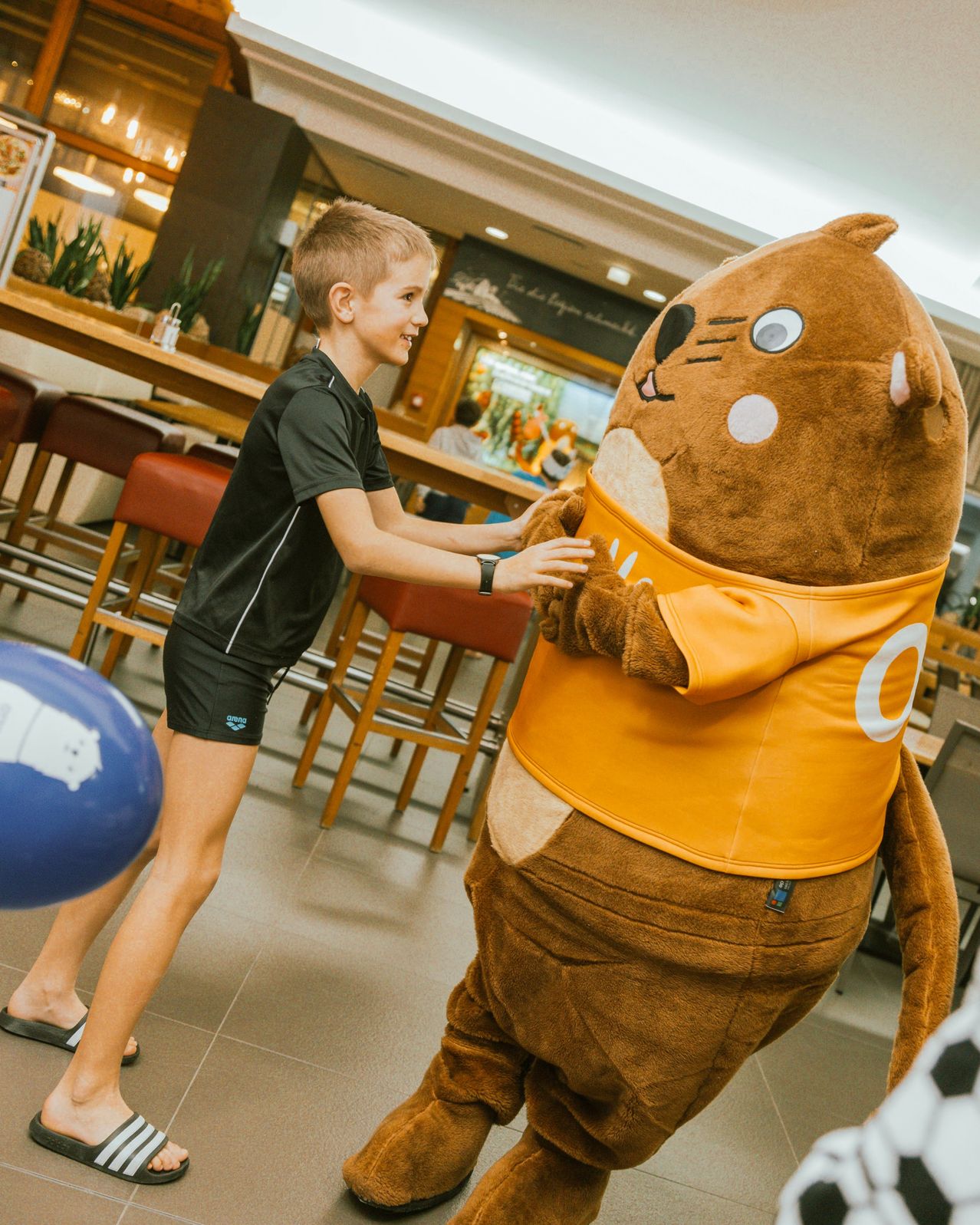 Ein junger Junge in schwarzer Sportbekleidung spielt mit einem Maskottchen in einer Cafeteria. Das Maskottchen, das ein orangefarbenes Shirt trägt, sieht freundlich aus. Der Junge hält einen blauen Ball.