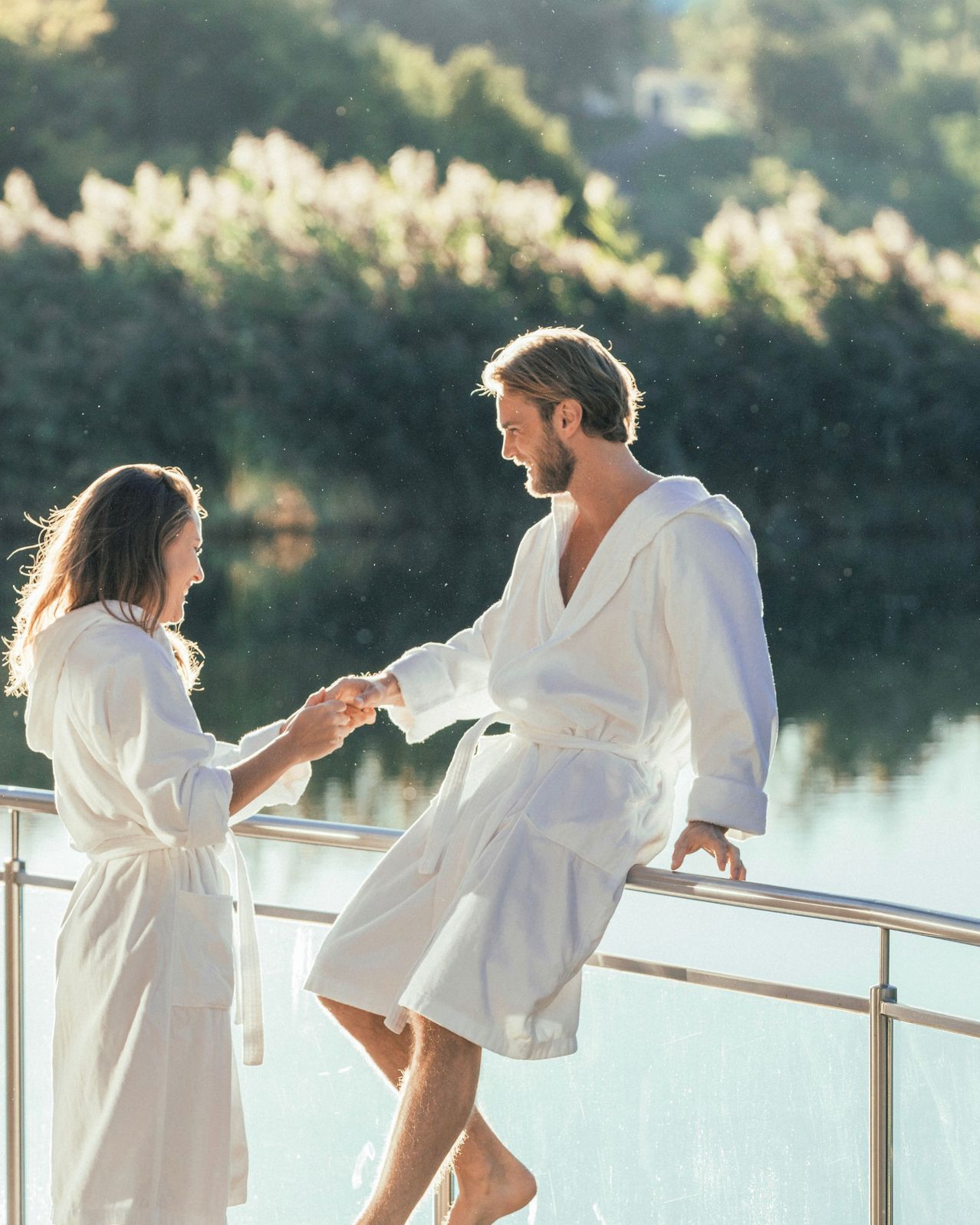 Ein Mann und eine Frau in weißen Gewändern halten sich an den Händen und stehen neben einem Geländer in der Nähe von Wasser. Der Mann lehnt sich über das Geländer.