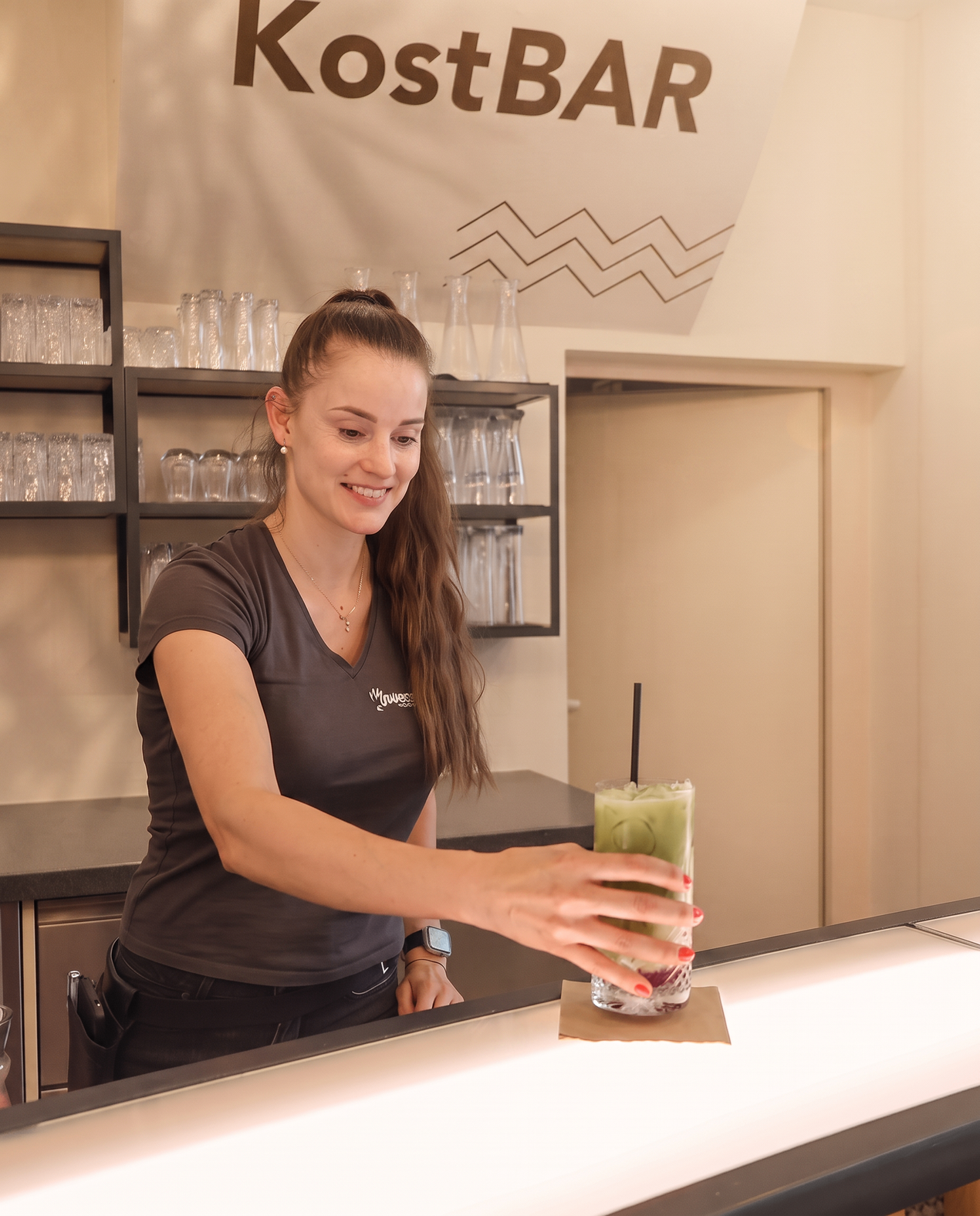 Eine Frau in einem schwarzen T-Shirt lächelt und hält einen grünen Drink in einer Bar.