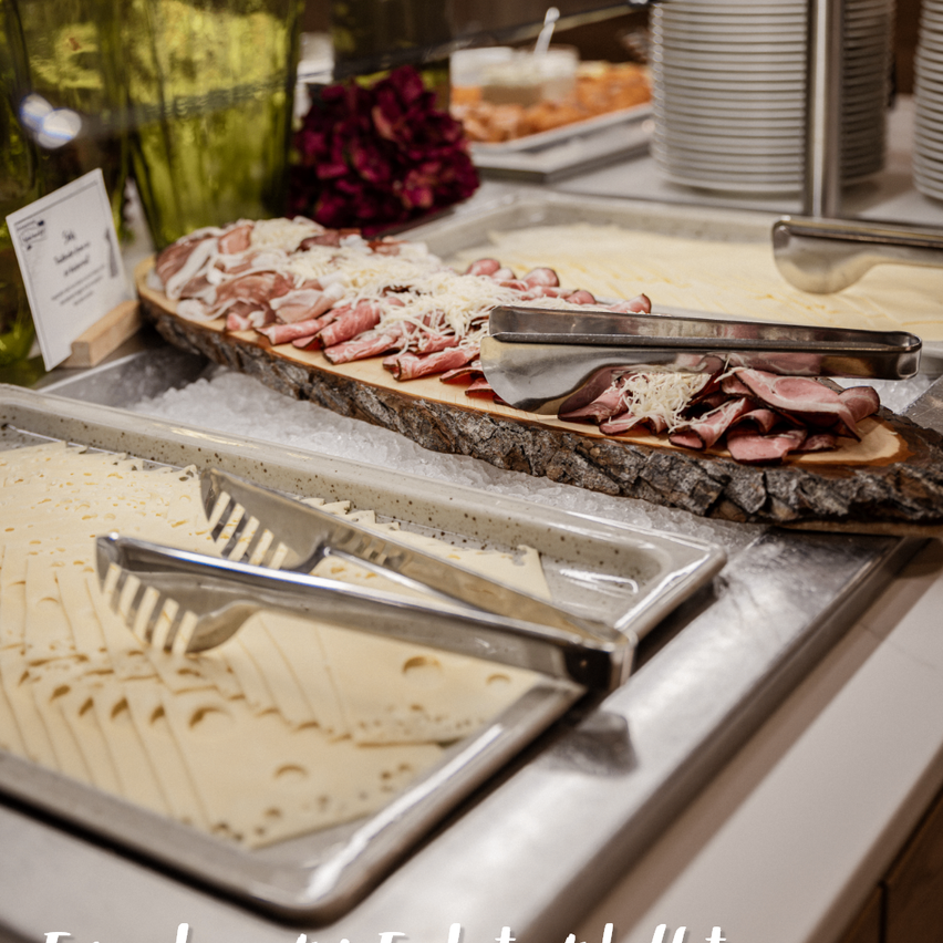 Ein Buffettisch mit einem großen, baumstammförmigen Tablett mit geschnittenem Fleisch und Käse, begleitet von einem Edelstahltablett mit Zangen und einem Tablett mit Käse.