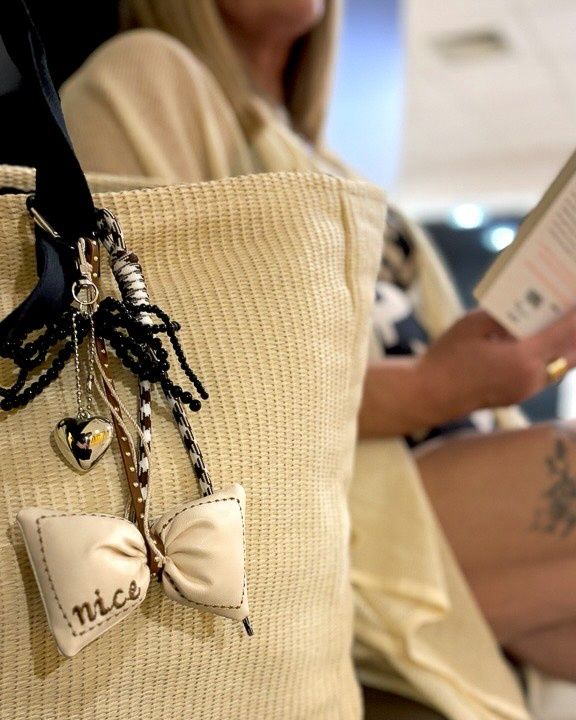 Eine beigefarbene Tragetasche mit dekorativen Anhängern, darunter ein Herz und eine Schleife, hängt an der Schulter einer Frau, während sie ein Buch liest.