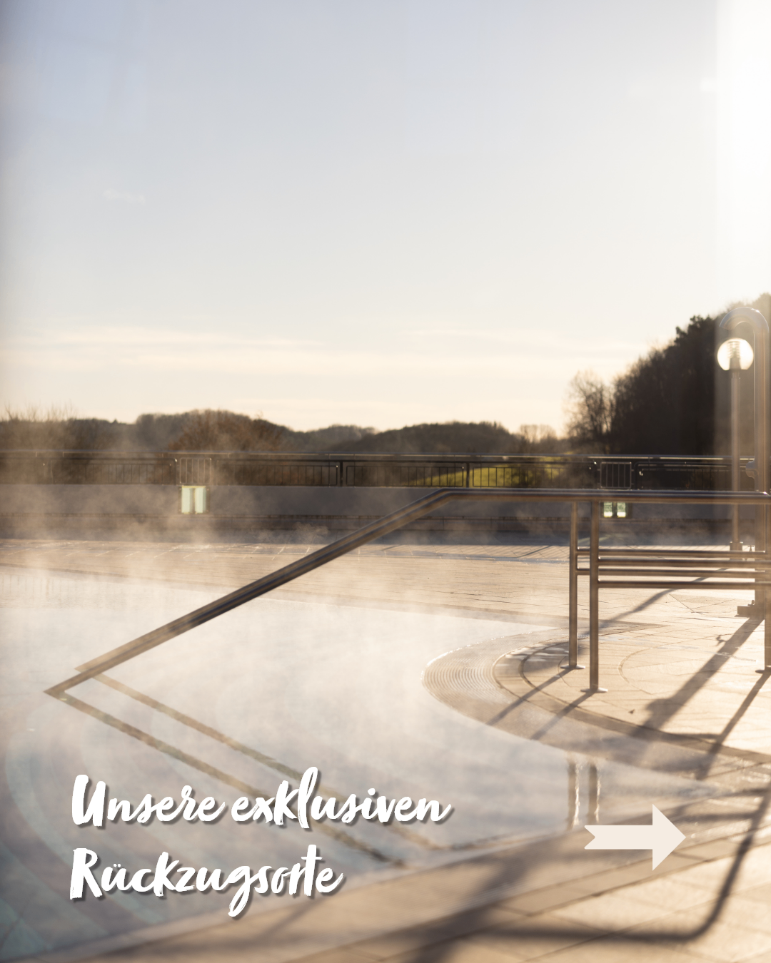 Ein Freibad mit aufsteigendem Dampf, umgeben von einem Metallgeländer. Im Hintergrund befindet sich eine ruhige Landschaft mit Bäumen und einem klaren Himmel. Das Bild hat einen Wasserzeichen mit dem Text 'Unsere exklusiven'.