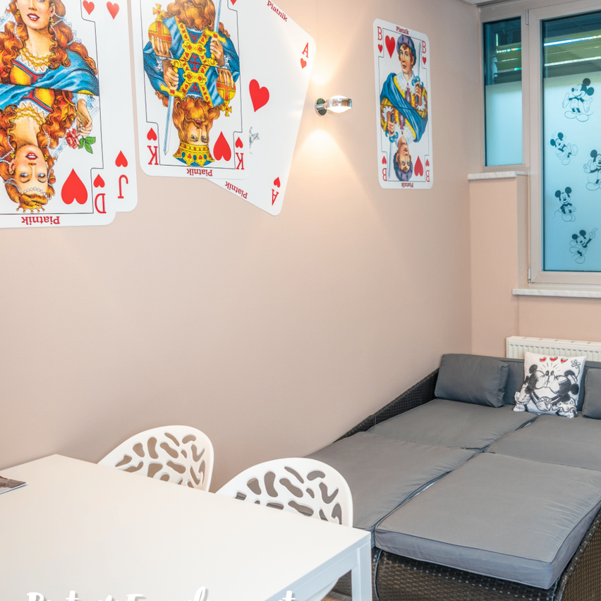 Ein Zimmer mit einem weißen Tisch, zwei Stühlen und einer Couch mit einem Micky-Maus-Kissen. Die Wand hat Spielkarten und eine Lampe.