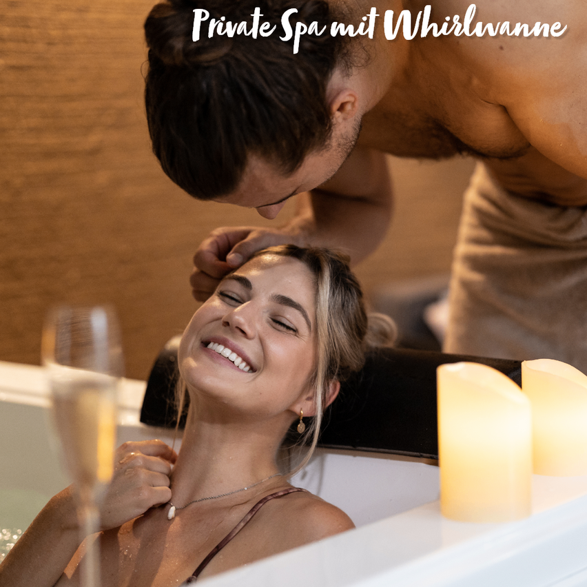Ein Mann beugt sich über eine lächelnde Frau in einer Badewanne, beide sehen entspannt aus, mit Kerzen und einem Glas Champagner in der Nähe.
