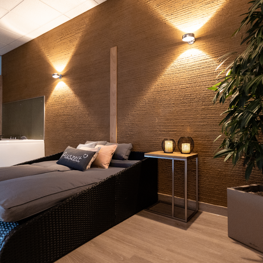 Ein gemütlicher Entspannungsbereich mit einem Weidenbett, Kissen und zwei brennenden Kerzen auf einem kleinen Tisch. Der Raum hat warmes Licht und eine Holzwand.