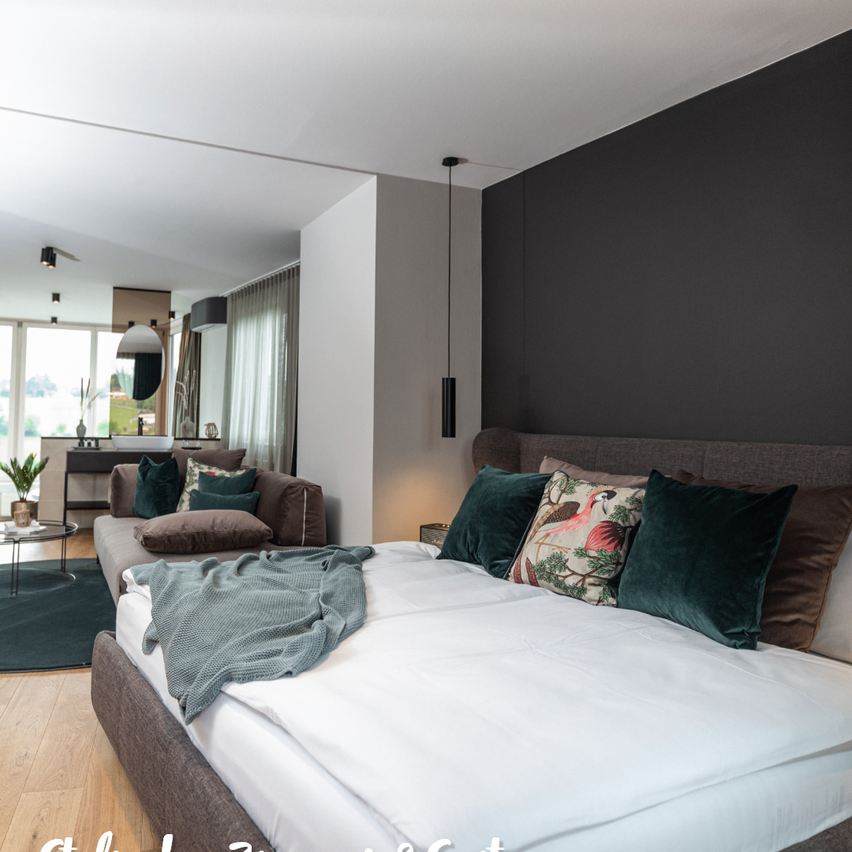 Ein modernes Schlafzimmer mit einem großen Bett, grauen Wänden und einem Holzboden. Das Bett hat weiße Laken und mehrere Kissen, darunter eines mit einem Flamingo-Design. Ein Sofa mit Kissen ist im Hintergrund.