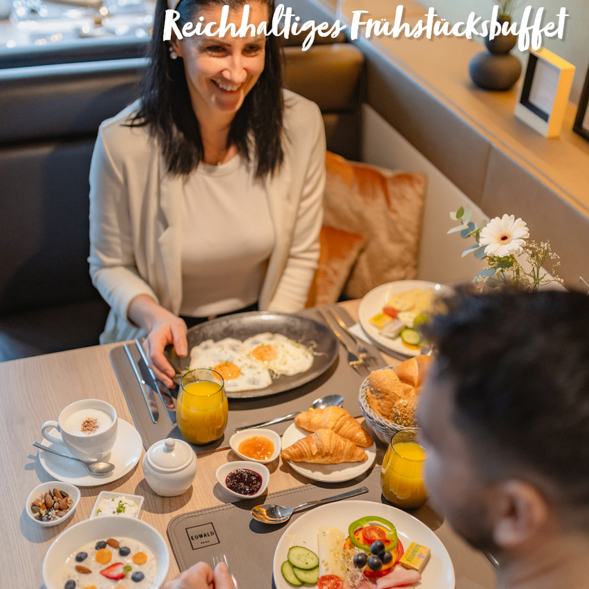 Eine lächelnde Frau und ein Mann genießen ein Frühstücksbuffet mit verschiedenen Speisen und Getränken auf einem Tisch.
