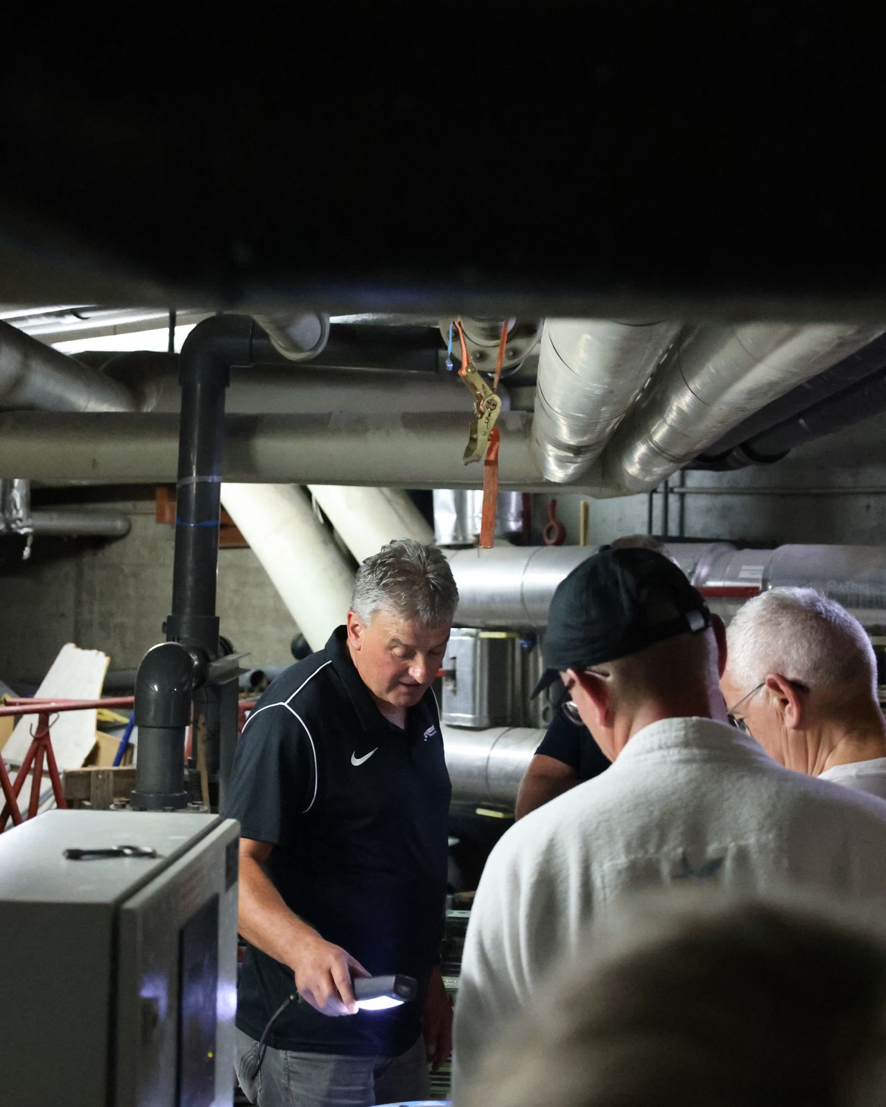 Three men are in a room with pipes and metal structures. The man in the middle is looking down at something.
