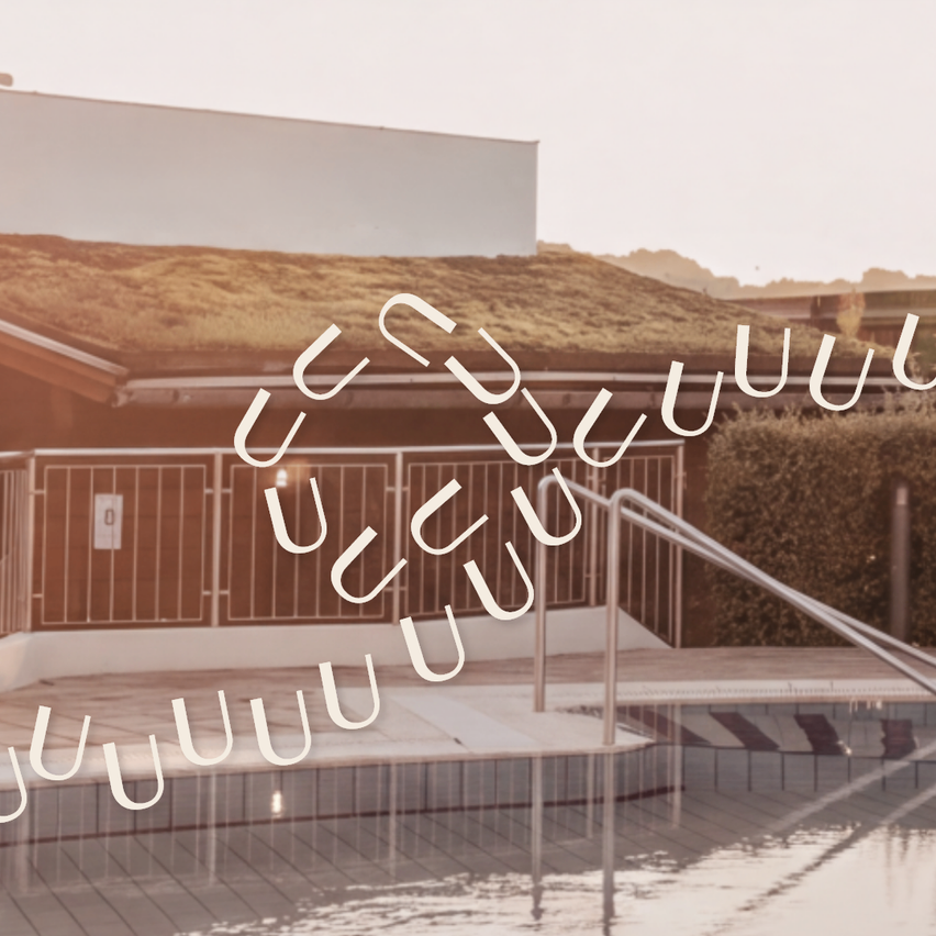 A modern building with a green roof and a swimming pool in front. The pool has a tiled floor and a metal handrail.