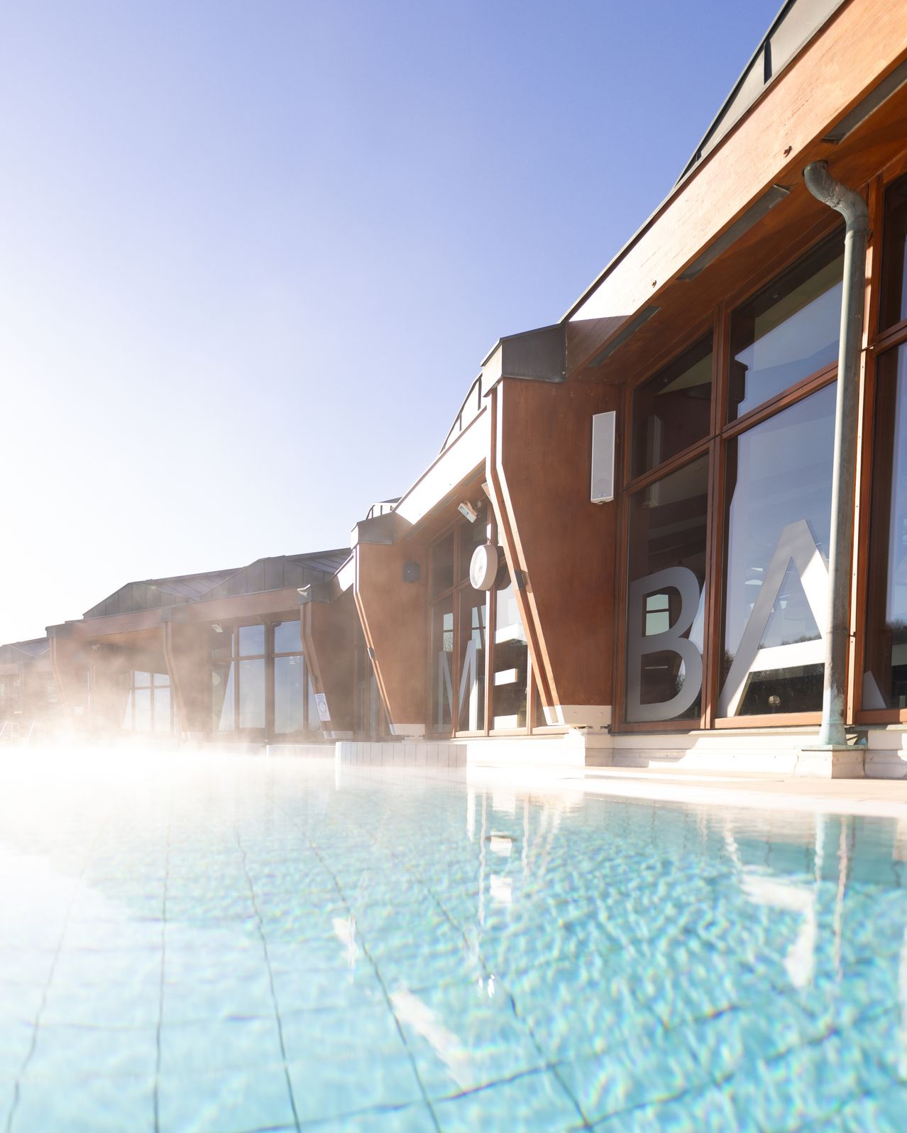 A modern building with a large pool in front, emitting steam. The building has large glass windows and wooden walls.