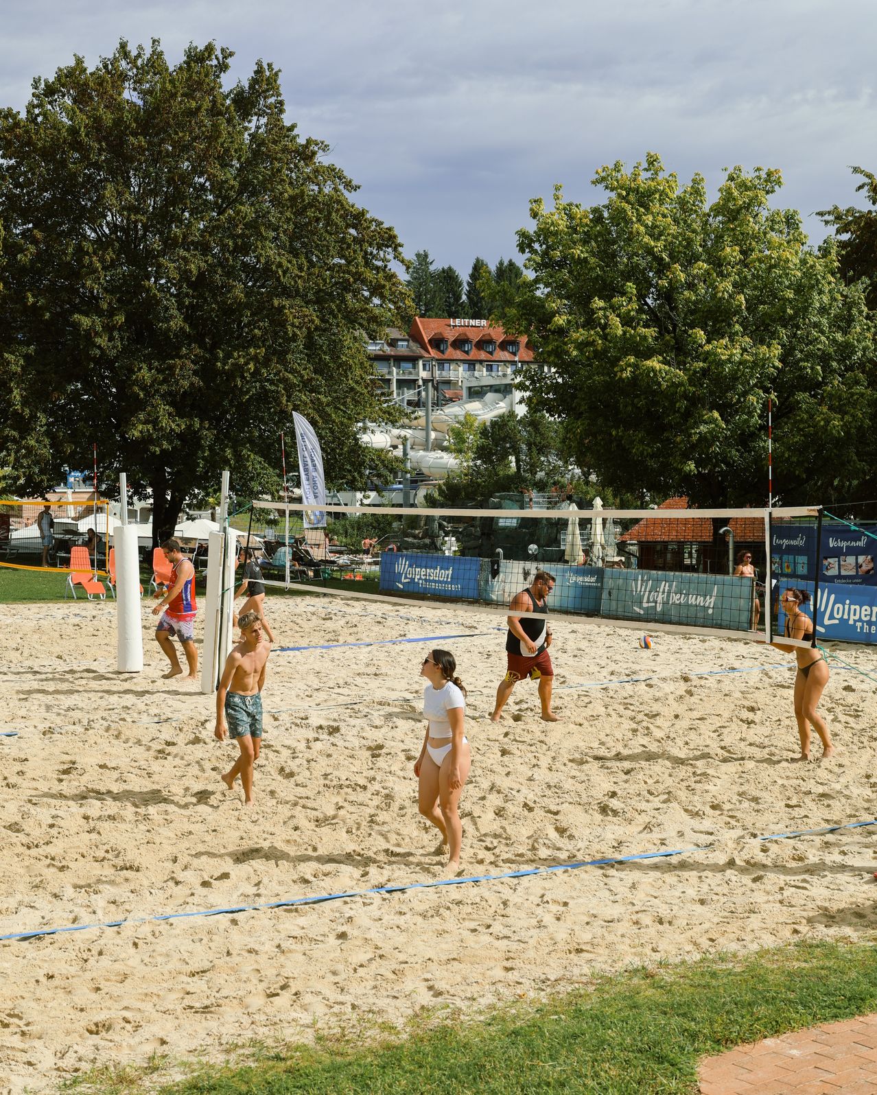 Beachvolleyball-Spieler am Sandplatz in Loipersdorf, Steiermark, mit Hotel im Hintergrund.