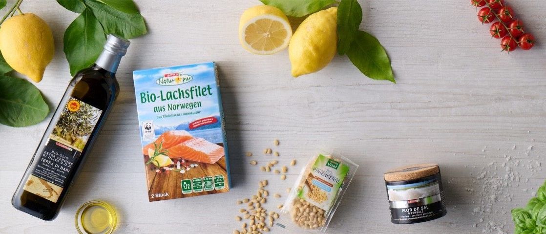 Auf einem weißen Tisch befinden sich verschiedene Lebensmittel, darunter Zitronen, Basilikum und Meersalz. Das Meersalz ist in einem Korkenglas.