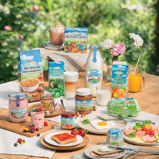 Ein Holztisch mit einer Auswahl an Bio-Lebensmitteln wie Granola, Joghurt, Kaffee, Milch, Eiern und frischem Obst. Es gibt auch verschiedene Gläser mit Aufstrichen und Cerealien. Im Hintergrund ist Grünery zu sehen.