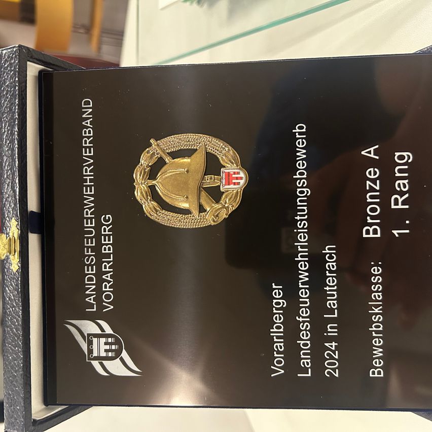 Eine schwarze Plakette mit einem goldenen Emblem und dem Text 'Landesfeuerwehrverband Vorarlberg' ist auf einer schwarzen Unterlage platziert. Die Plakette befindet sich in einer Schachtel.