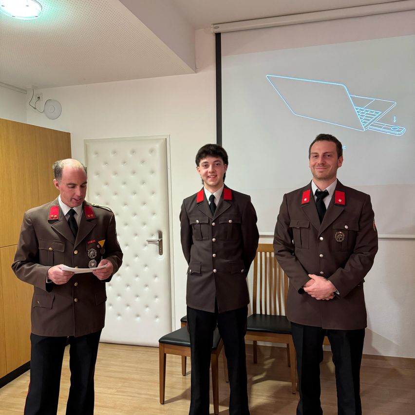 Drei Männer in Militäruniformen stehen nebeneinander, einer hält ein Papier und die anderen beiden lächeln. Dahinter befindet sich eine Tür mit einem weißen Kissen und einem Holzstuhl. An der Wand ist eine Projektionsleinwand mit einem Laptop-Diagramm.