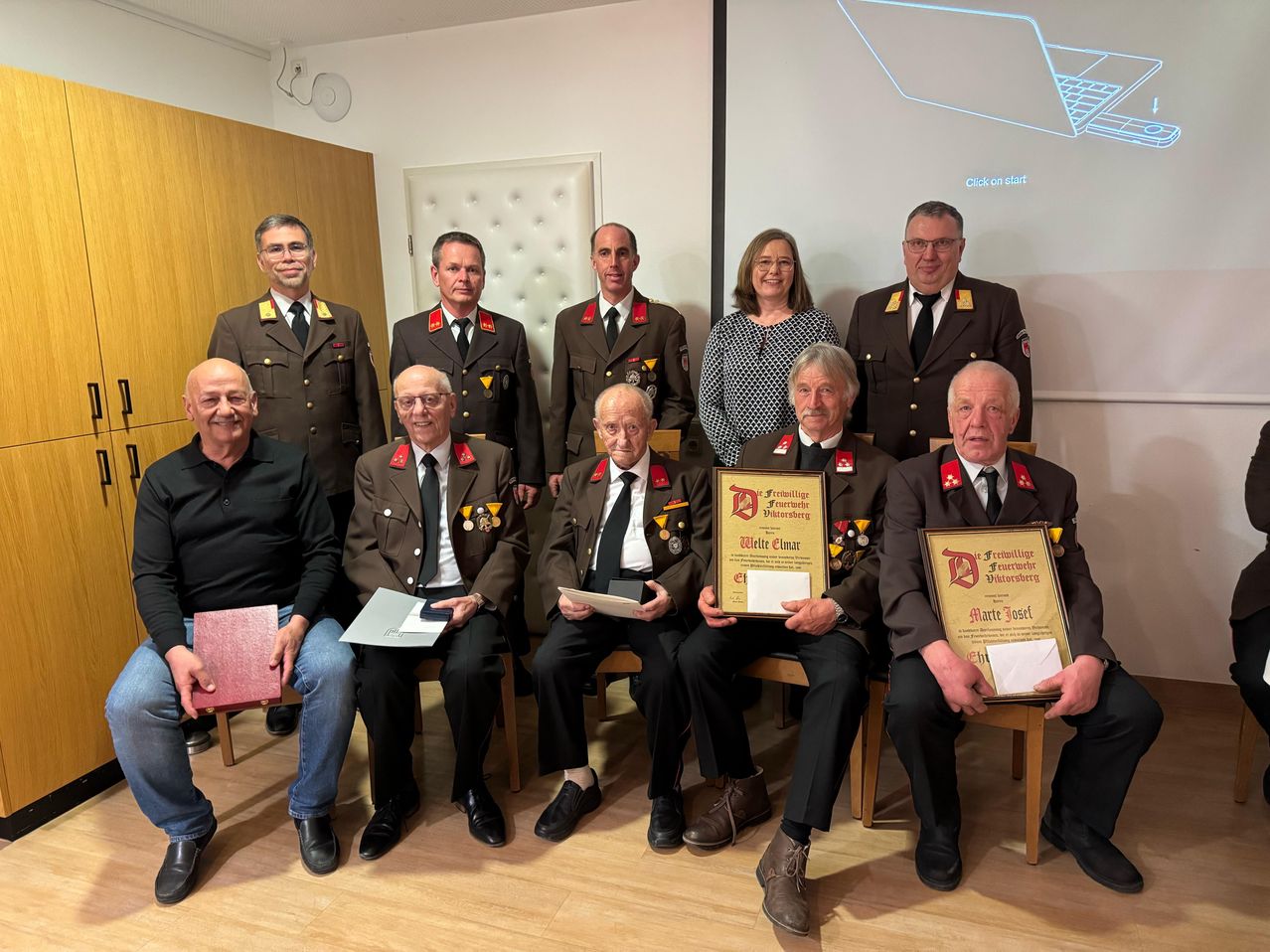 Eine Gruppe von Männern und Frauen, einige in Uniform, sitzen und stehen in einem Raum. Ein Mann in der vorderen Reihe hält ein Zertifikat.