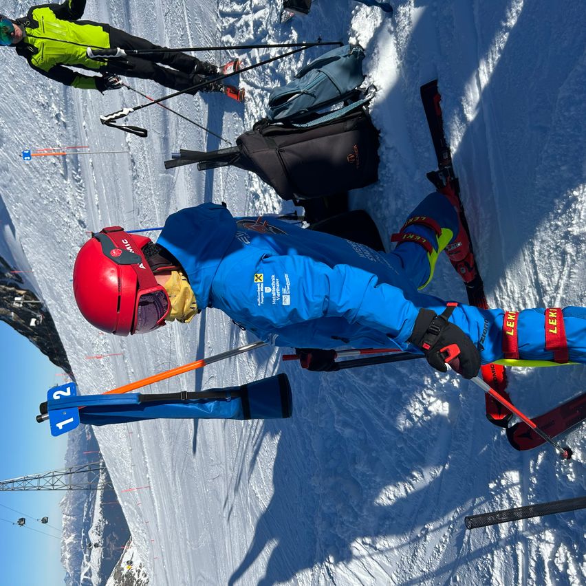 Ein Skifahrer in blauer Ausrüstung liegt auf dem Schnee und hält einen Skistock. In der Nähe steht ein anderer Skifahrer in grüner und schwarzer Ausrüstung mit Skiern und Stöcken. Taschen und Skier sind auf dem Schnee verstreut.
