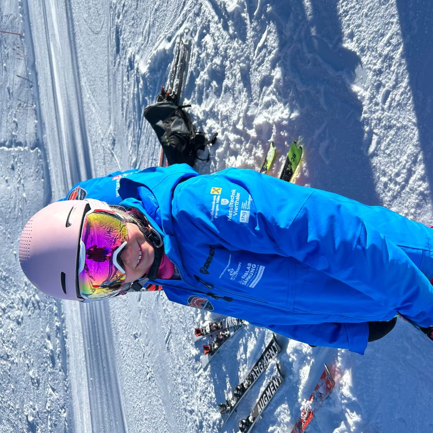 Ein Skifahrer liegt auf einem schneebedeckten Hang mit Skiern und Ausrüstung herum, trägt eine blaue Jacke und einen rosa Helm und lächelt.