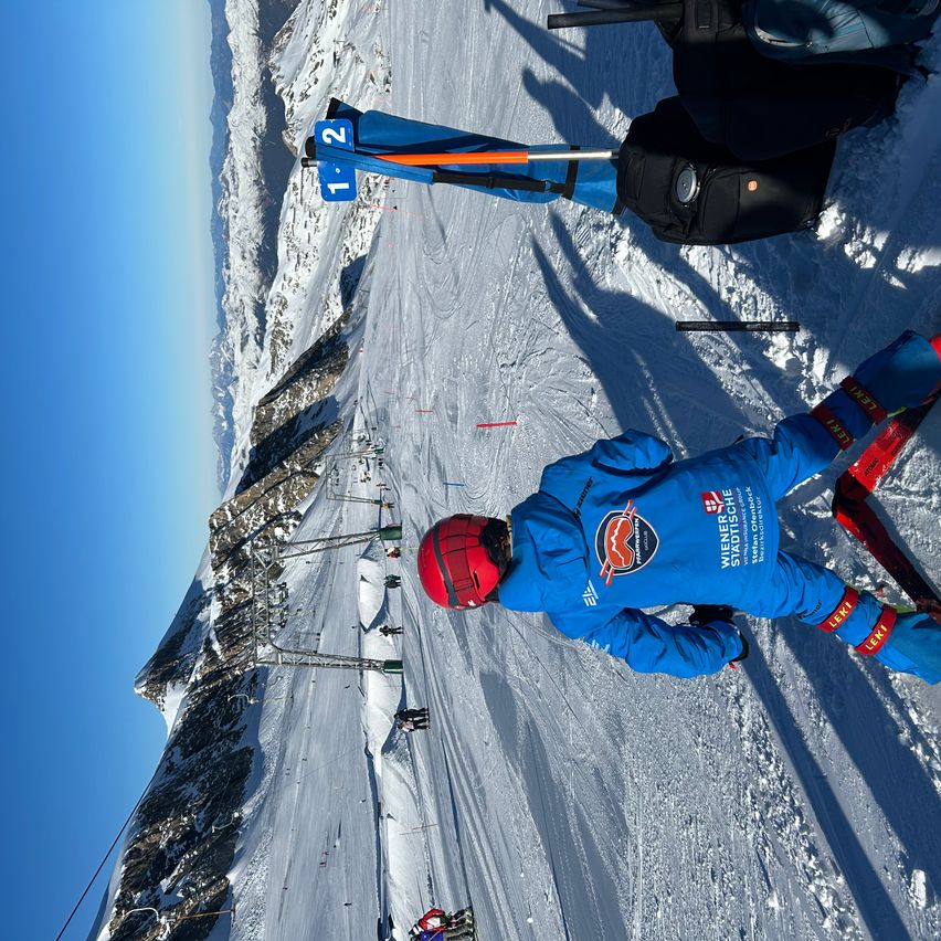Ein Skifahrer in einem blauen Anzug und einem roten Helm liegt auf der schneebedeckten Piste. Die Piste hat ein Schild mit der Nummer 12, und eine rote und orange Fahne ist am Pfahl befestigt. Der Himmel ist blau und der Berg hat eine felsige Oberfläche.