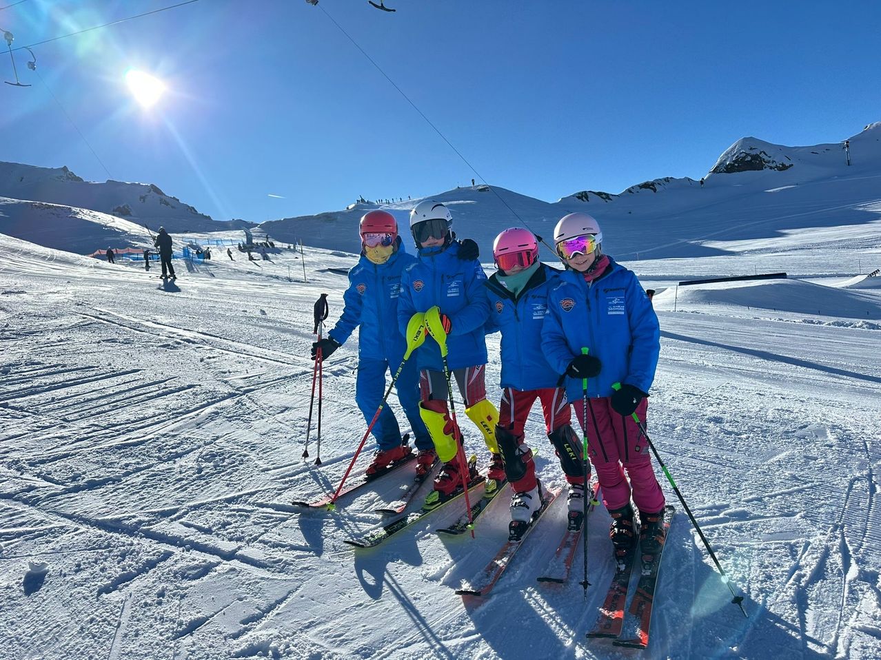 Vier Skifahrer, gekleidet in blaue und rote Skianzüge, stehen auf einem verschneiten Berg. Sie tragen alle Helme und halten Skistöcke. Hinter ihnen ist der Berg mit Schnee bedeckt, und in der Ferne fahren Menschen Ski.